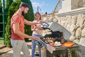 Un homme barbu fait griller des saucisses et des légumes sur un barbecue à gaz en acier inoxydable dans une cuisine d’été en plein air, pendant qu’une femme tient un verre de vin rouge et le regarde